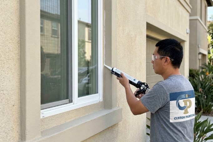 Painter running a bead of caulk where a white window frame meets a beige stucco wall