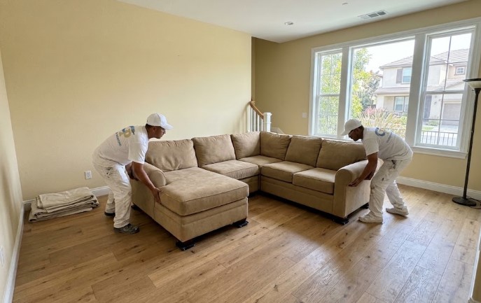 Two painters in white lifting a tan sectional sofa away from the wall of a living room, with drop cloths folded nearby
