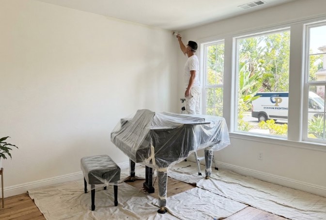 Grand piano and bench wrapped in plastic in place on drop cloths while a painter cuts in along the ceiling