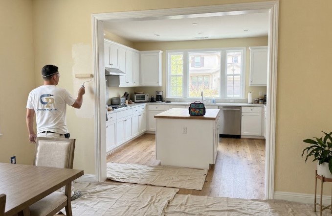 Painter rolling a dining room wall over drop cloths while the open kitchen next to it stays in use